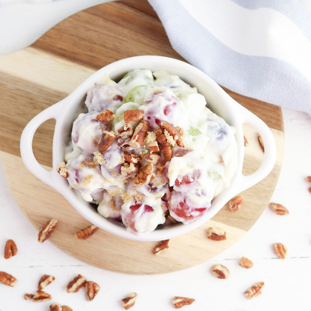 Creamy Grape Salad topped with pecans in white bowl on wooden serving board.