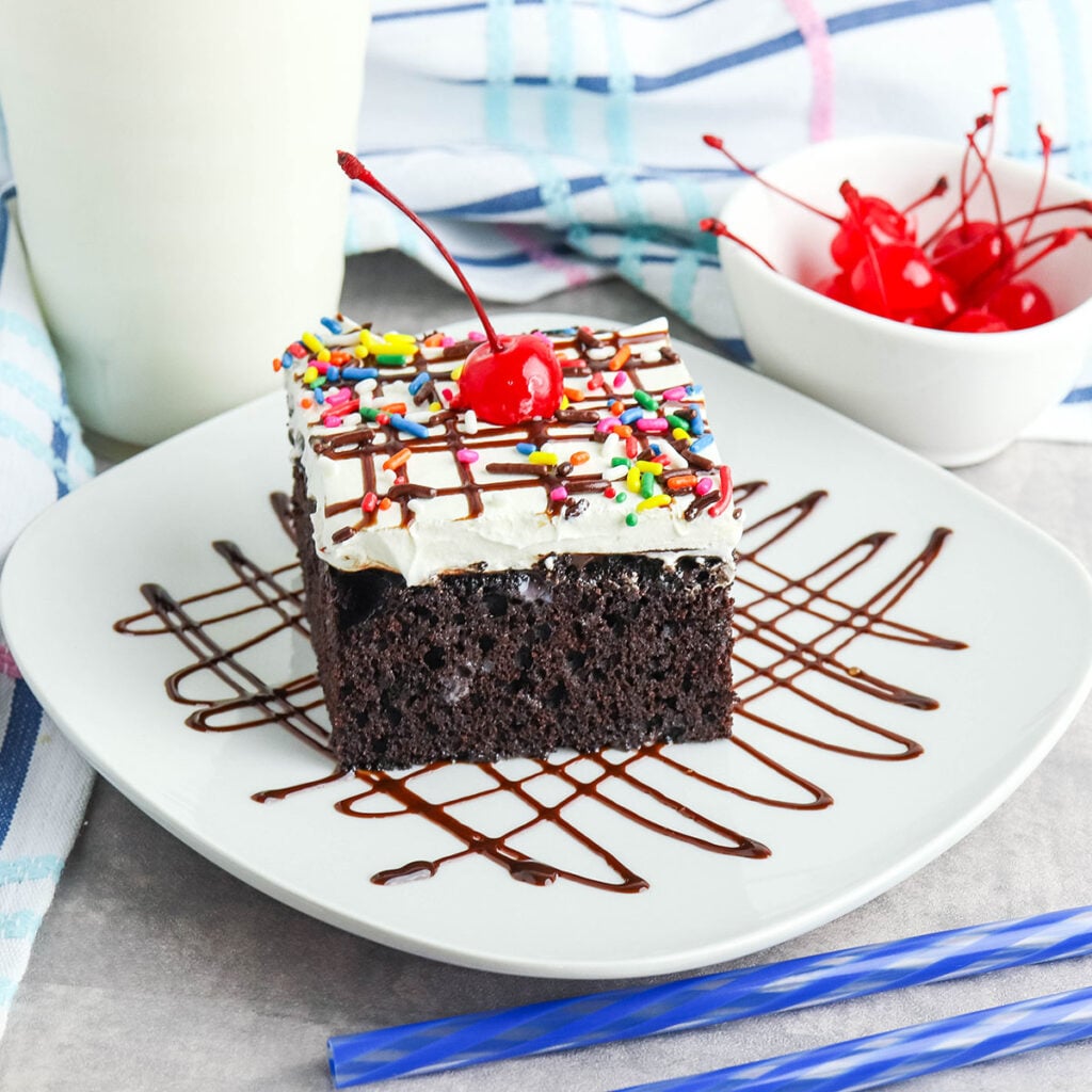 Slice of Hot Fudge Poke Cake on white plate drizzled with chocolate and topped with cherry.