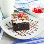 Slice of Hot Fudge Poke Cake on white plate drizzled with chocolate and topped with cherry.