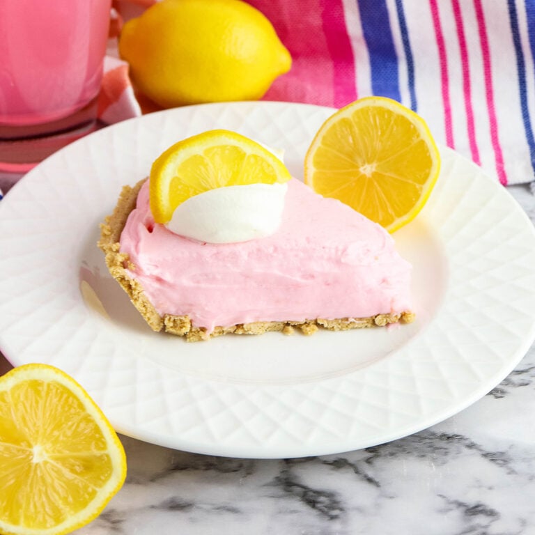 Slice of Pink Lemonade Pie on white plate topped with cool whip and lemon wedge.