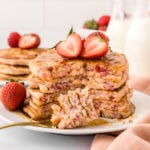 A stack of strawberry pancakes drizzled with maple syrup on a plate with a bite on a fork.