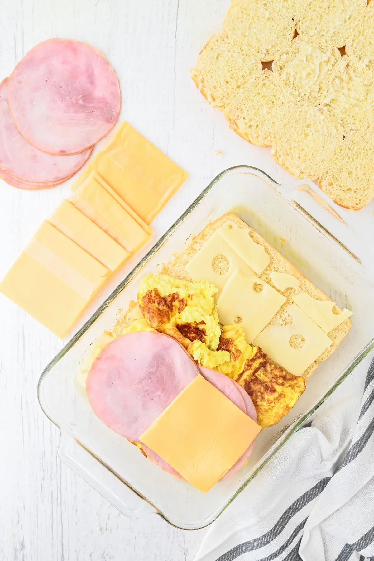 Layering cheese, eggs, and ham over the bottom of the Hawaiian rolls in a glass baking dish.
