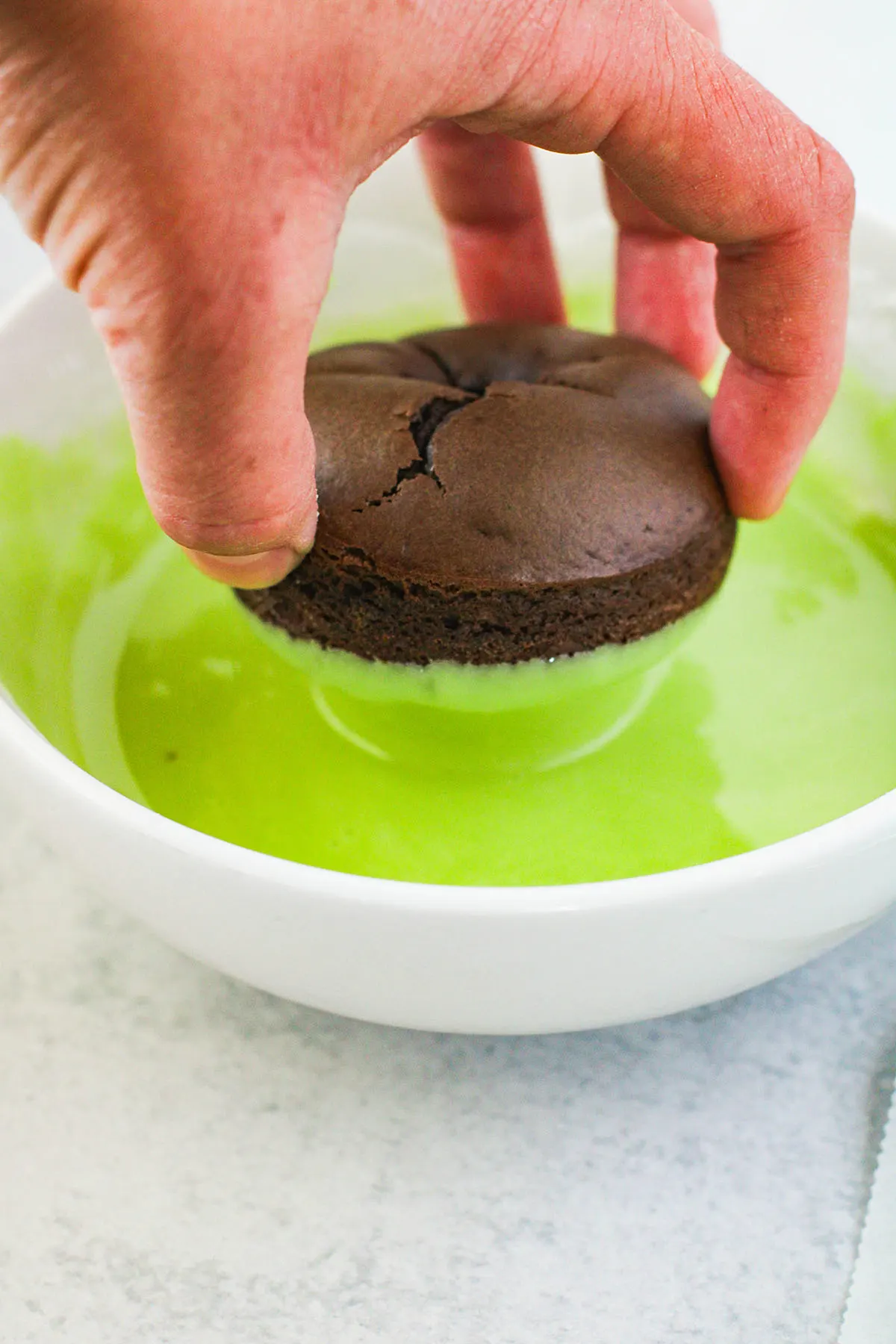 Dipping chocolate donut in green glaze in a bowl.