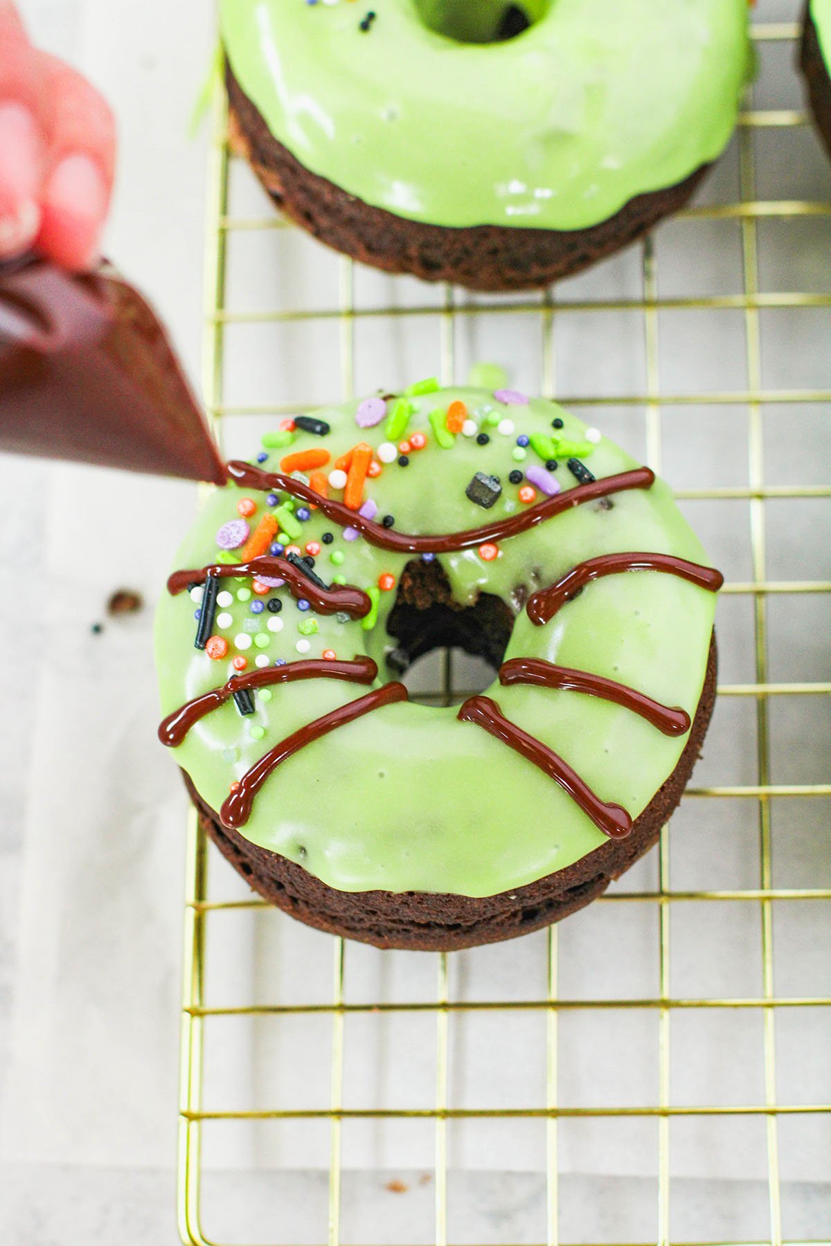 Piping chocolate glazed spider legs to a green glazed chocolate donut.