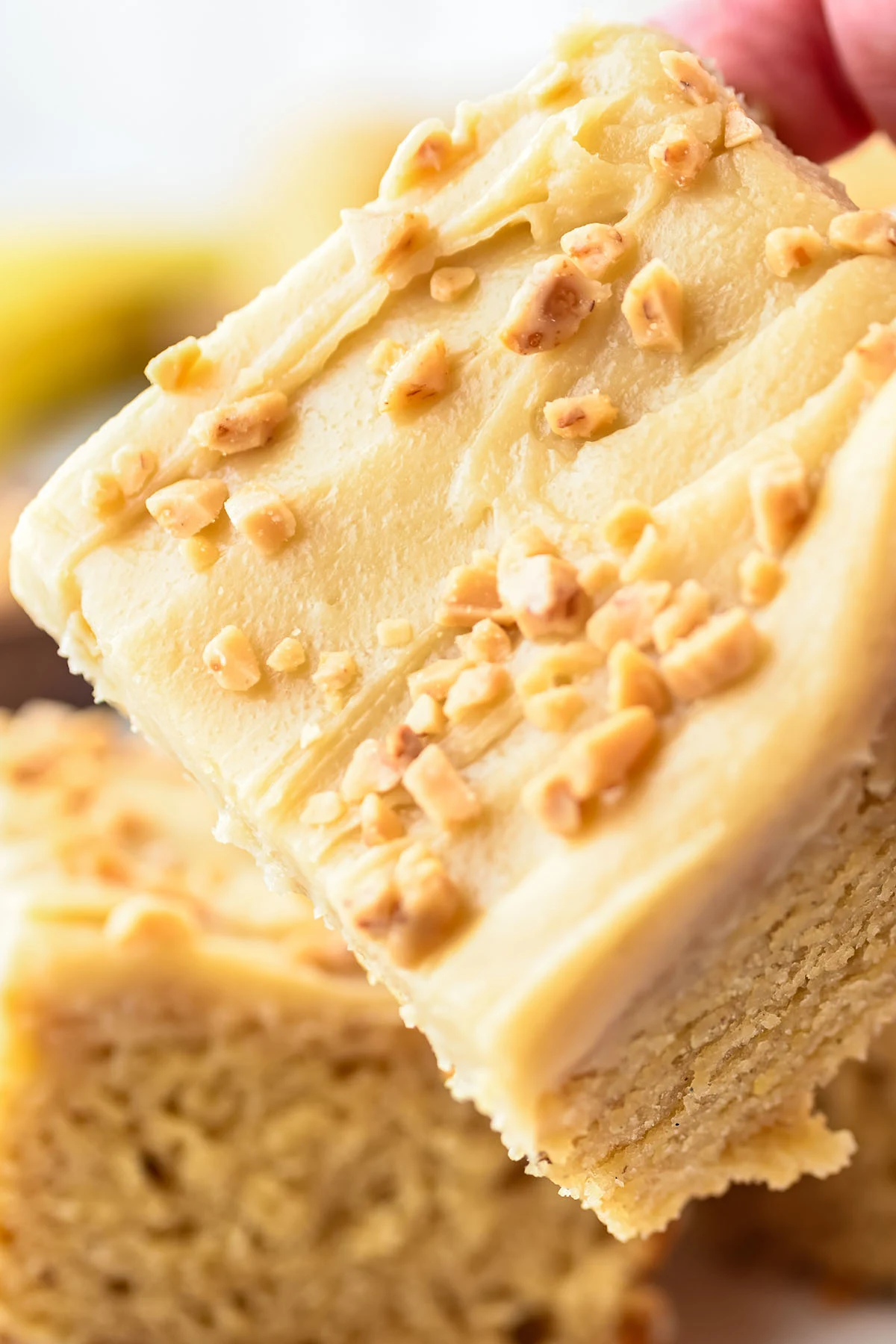 A hand holding a slice of banana blondies with brown butter frosting topped with chopped nuts.