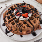 Two chocolate waffles on a white plate topped with whipped cream, strawberries, and chocolate syrup.