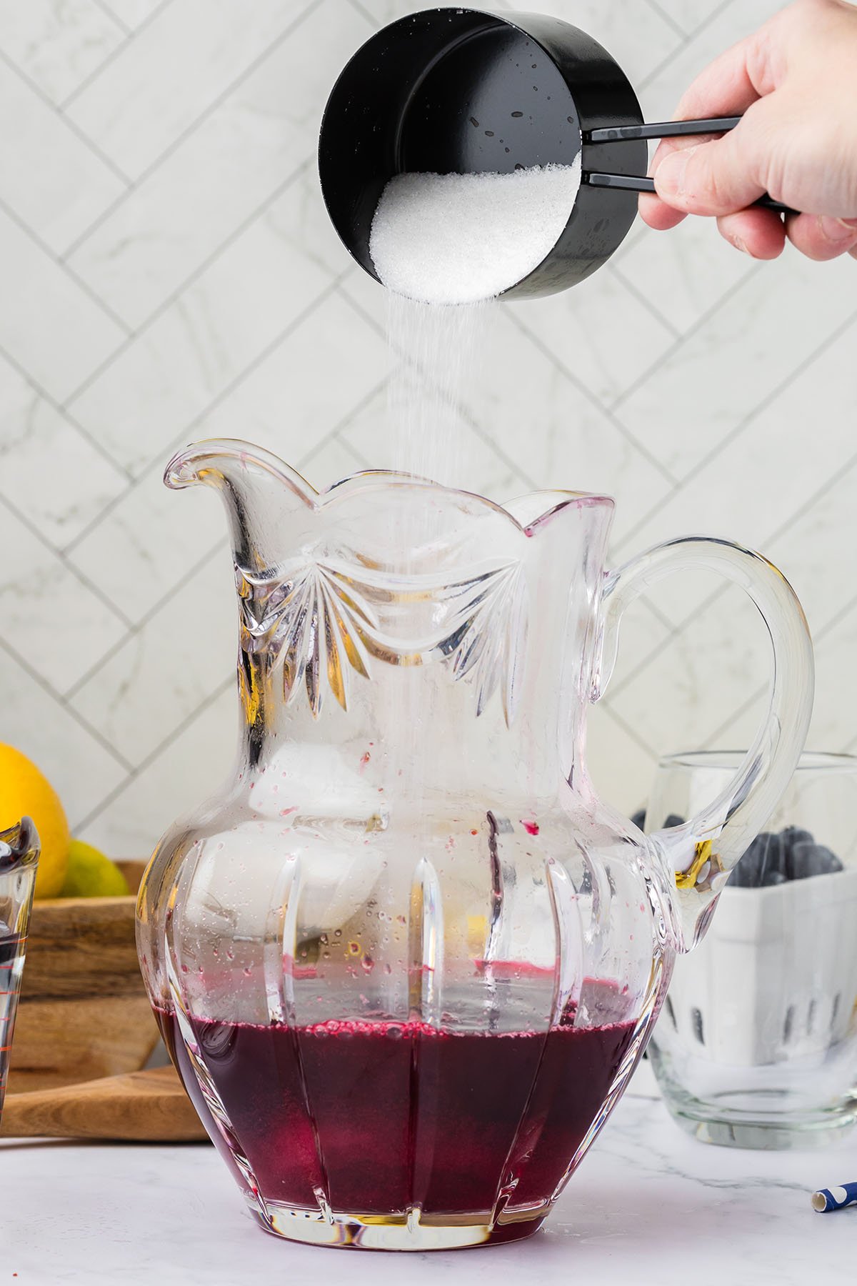Sugar is added to the blueberry lemonade liquid in the glass pitcher.