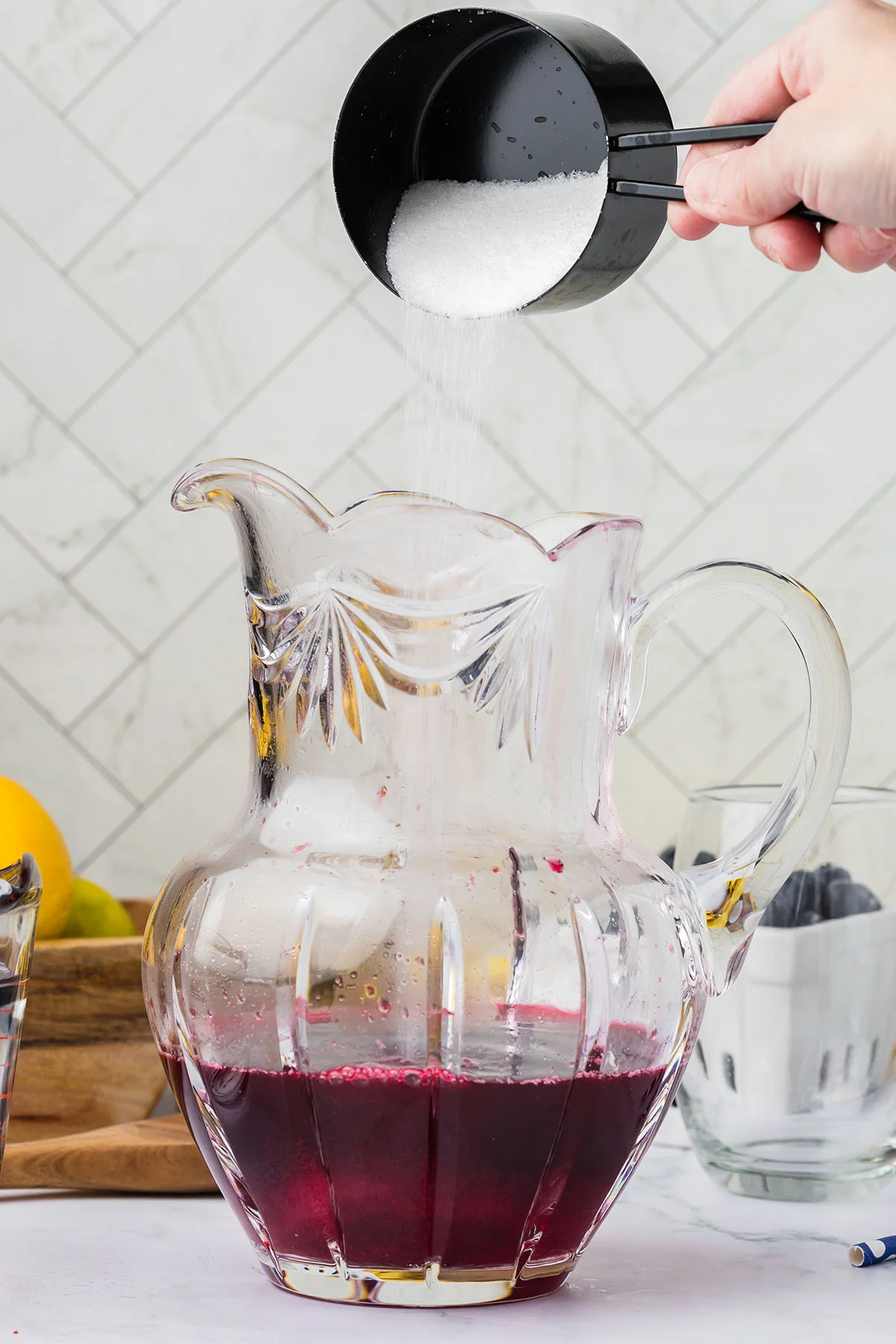 Sugar is added to the blueberry lemonade liquid in the glass pitcher.