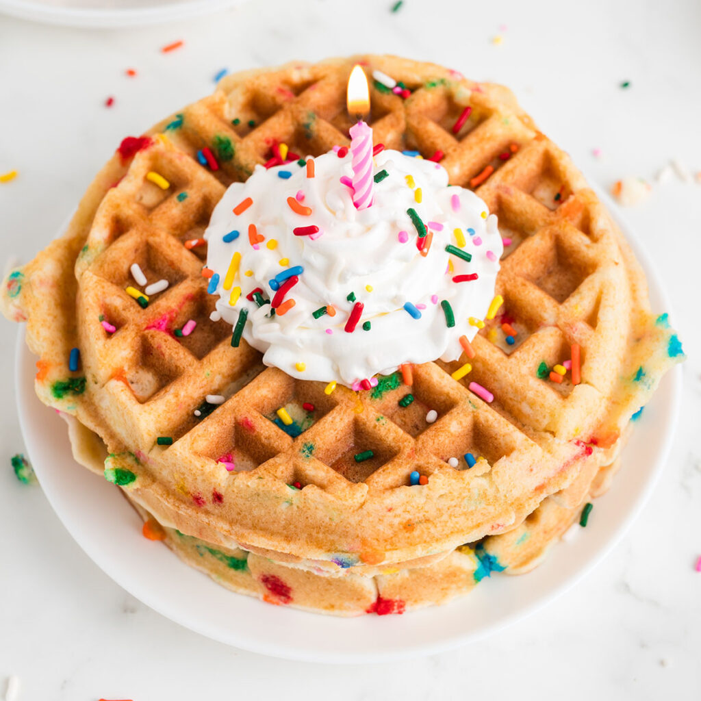 Birthday Cake Waffles on a plate topped with whipped cream, sprinkles, and a candle.