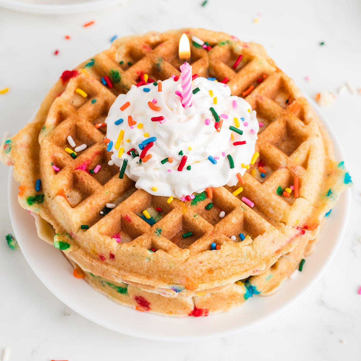Birthday Cake Waffles on a plate topped with whipped cream, sprinkles, and a candle.