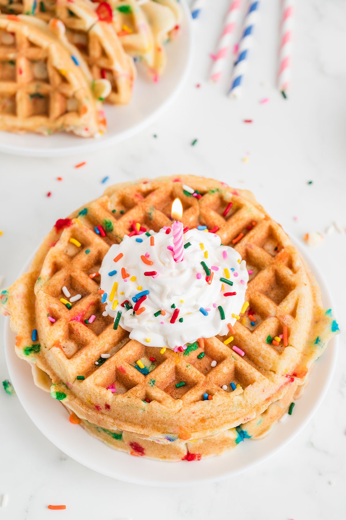 Birthday Cake Waffles topped with whipped cream, sprinkles, and a candle on a plate.