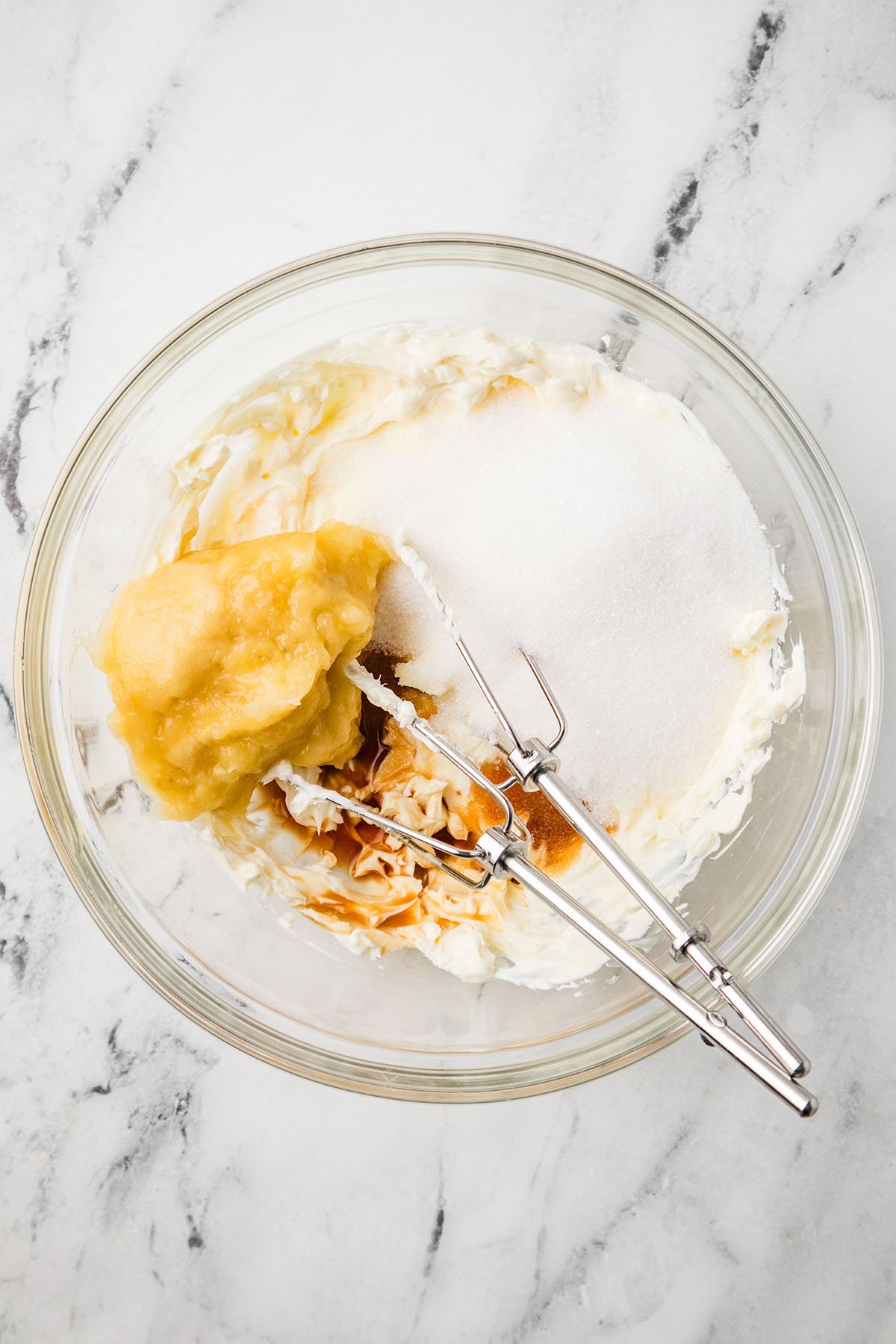 Sugar, vanilla, and mashed banana are mixed into the cream cheese in a glass mixing bowl.