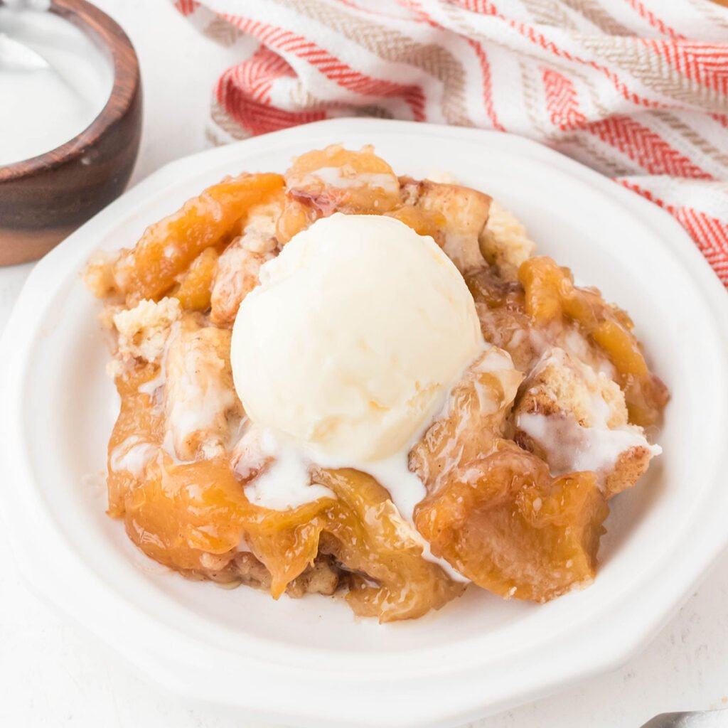 A serving of Cinnamon Roll Peach Cobbler in a white bowl with a scoop of vanilla ice cream.