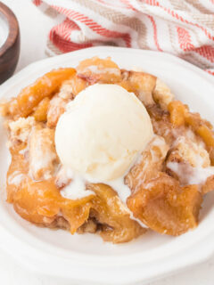 A serving of Cinnamon Roll Peach Cobbler in a white bowl with a scoop of vanilla ice cream.