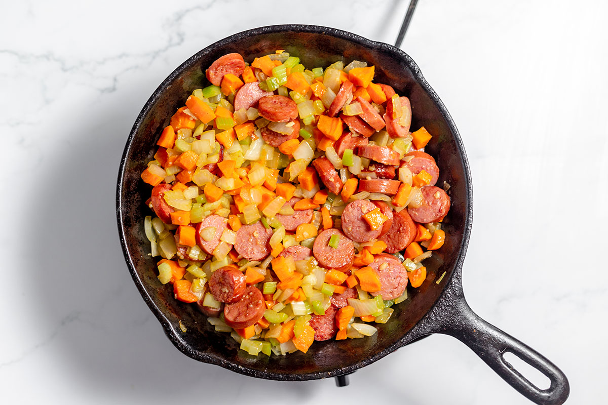 Saut&eacute;ed celery, onion, carrots, and smoked sausage in a cast-iron skillet.