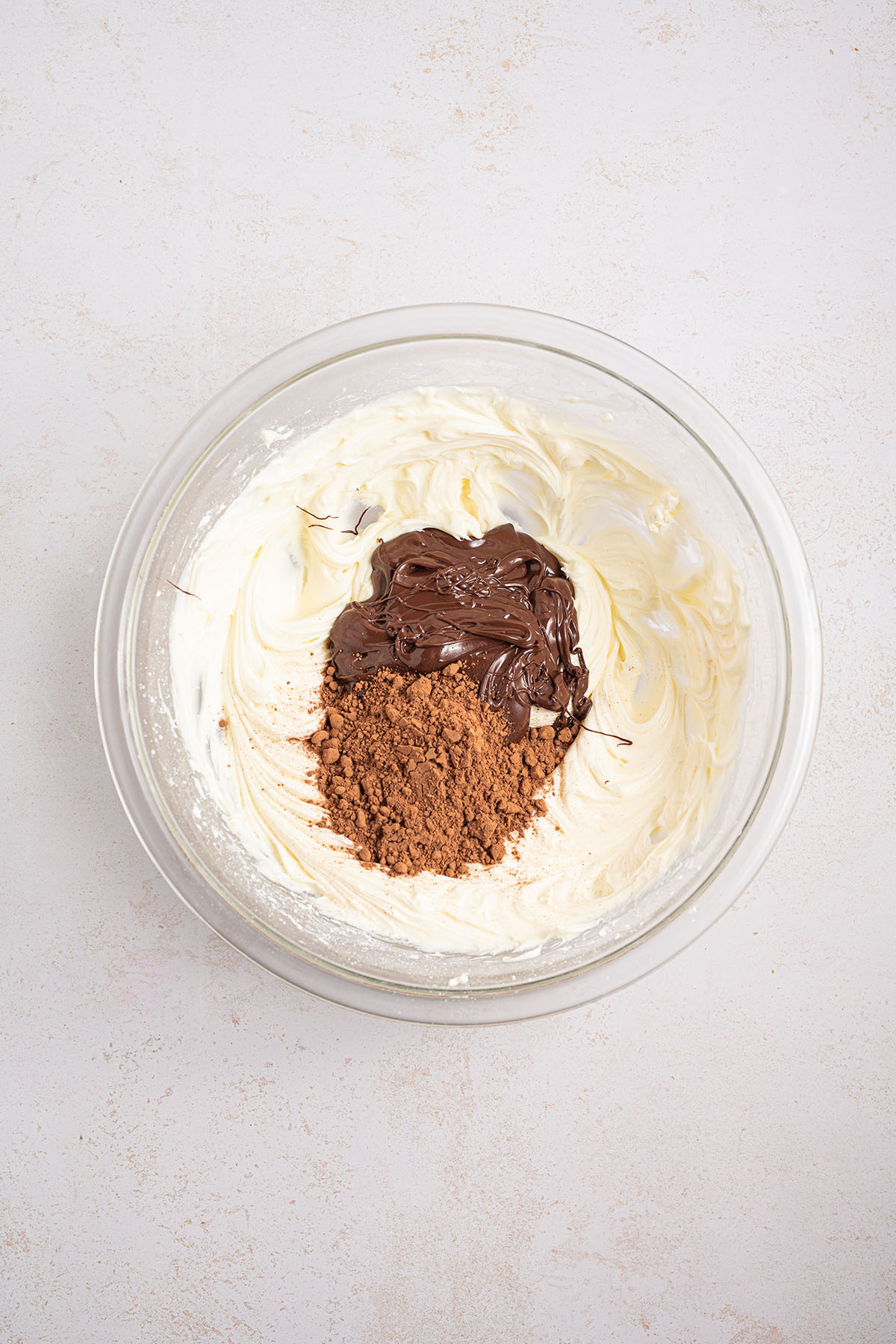 Melted chocolate and cocoa powder added to the cream cheese mixture in the glass mixing bowl.